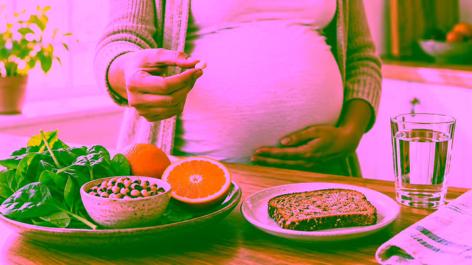 A prenatal vitamin beside spinach, beans, citrus, and a glass of water on a calm kitchen table