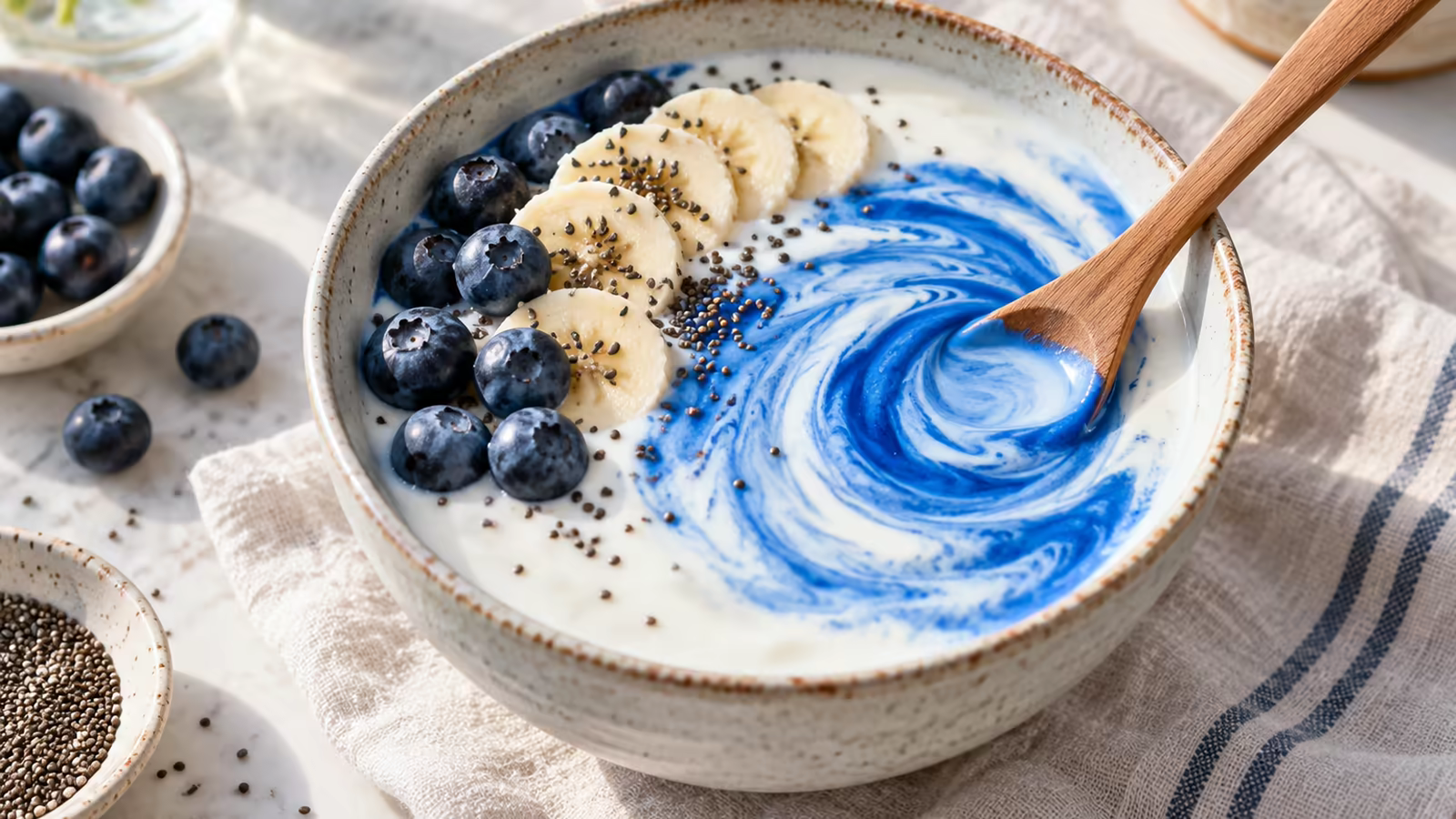 A spoon swirling blue spirulina into a pale smoothie bowl with berries, chia seeds, and yogurt