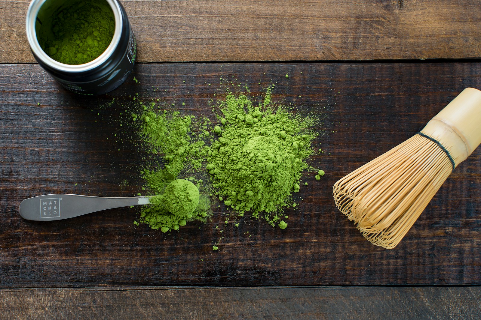 Matcha powder, a spoon, and a bamboo whisk on a wooden surface