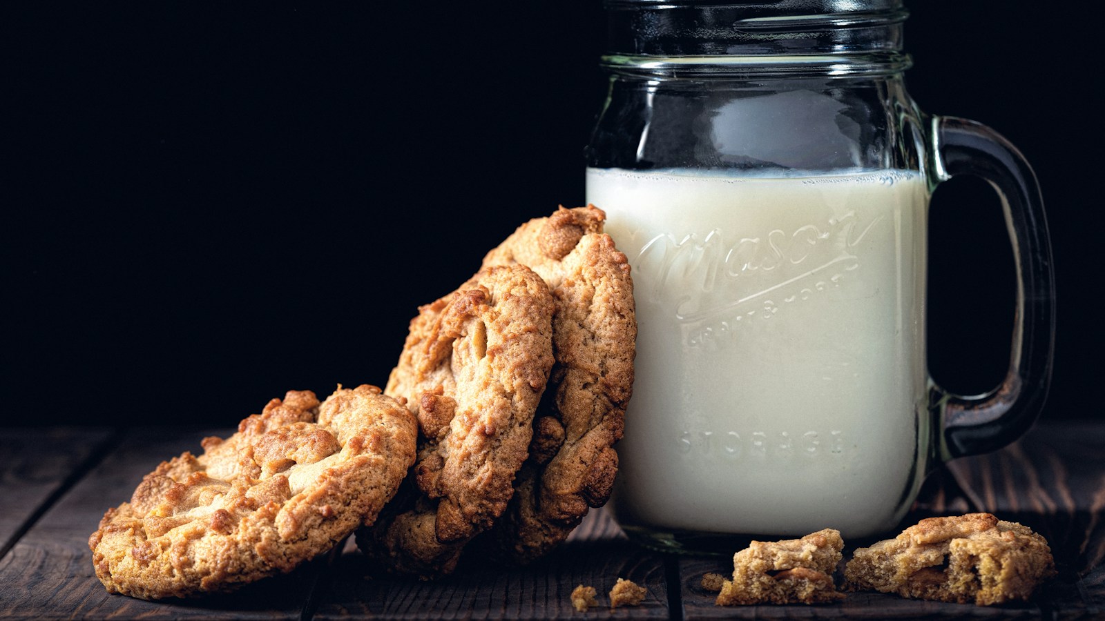 A glass mug of milk beside cookies on a dark table